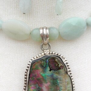 Puede incluir: Juego de collar y pendientes. El collar presenta un colgante con una concha de abul&oacute;n iridiscente, engastada en un marco plateado. El collar tiene cuentas verde p&aacute;lido, y los pendientes tienen cuentas ovaladas oscuras y verde claro.