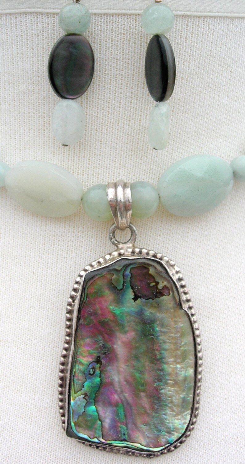 Puede incluir: Juego de collar y pendientes. El collar presenta un colgante con una concha de abul&oacute;n iridiscente, engastada en un marco plateado. El collar tiene cuentas verde p&aacute;lido, y los pendientes tienen cuentas ovaladas oscuras y verde claro.