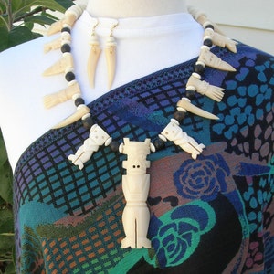 May include: A white bone necklace with black beads and carved bone figures. The necklace is worn on a mannequin with a patterned shirt. The necklace includes a pendant with a carved bone figure.