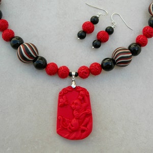 Puede incluir: Un collar de cuentas rojas y negras con un colgante rojo con un diseño tallado. El colgante está unido al collar con una cadena de plata. El collar se combina con pendientes a juego.