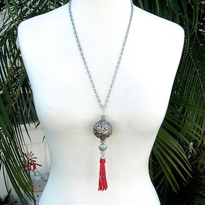 Puede incluir: Un collar de cadena de plata con un gran colgante de plata adornado y una borla roja. El colgante es redondo y tiene un diseño detallado.