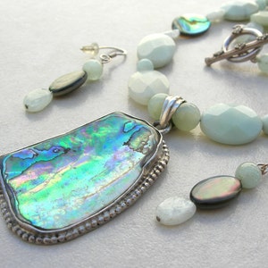 Puede incluir: Un conjunto de joyas con collar, colgante y pendientes. El collar y los pendientes tienen cuentas de color verde claro e iridiscentes. El colgante tiene forma rectangular con borde plateado y centro de concha de abul&oacute;n iridiscente.