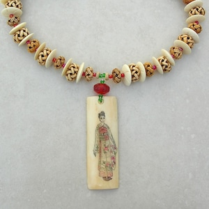 May include: A beige and brown woven bead necklace with a red and green bead accent. The necklace has a pendant featuring a hand-painted image of a woman in a traditional Japanese kimono.
