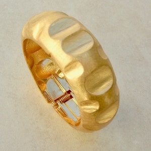 Puede incluir: Un brazalete dorado con un diseño texturizado y acanalado. El brazalete tiene un cierre con bisagra.