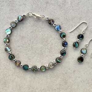 Puede incluir: Un conjunto de joyería compuesto por una pulsera y un par de pendientes colgantes. La pulsera presenta cuentas redondas de concha de abulón iridiscente que se alternan con pequeños espaciadores metálicos cuadrados de tono plateado.