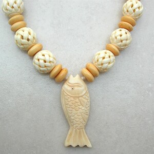 May include: A white bone necklace with a carved fish pendant. The necklace is made of round beads with a woven pattern.