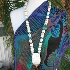 May include: A long, handcrafted necklace with large, off-white, rectangular and oval beads, with smaller turquoise and brown beads. The necklace has a large, white, rectangular pendant. The necklace is displayed on a mannequin, draped over a patterned shawl.