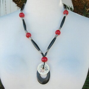 May include: A black and red beaded necklace with a large, oval-shaped pendant. The pendant is made of white bone with a floral design and a black bone ring.