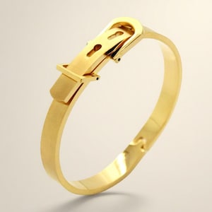 May include: A gold-colored bracelet with a buckle design. The bracelet features a rectangular buckle with a keyhole detail and a rounded band. The metal has a matte finish, and the overall design is minimalist and elegant.