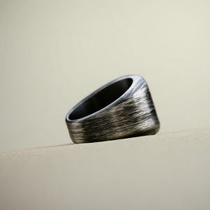 May include: A silver-toned ring with a brushed metal texture. The ring has a thick band and a flat, rectangular top. The interior of the ring is dark, creating a contrast with the exterior.
