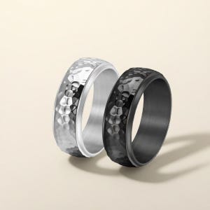 Könnte beinhalten: Zwei Ringe mit einem strukturierten, gehämmerten Metalldesign. Ein Ring ist silberfarben, der andere schwarz. Die Ringe haben einen modernen, minimalistischen Stil. Die Ringe sind wahrscheinlich aus Edelstahl oder einem ähnlichen Metall gefertigt.