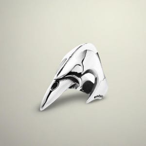 May include: A polished silver ring with a bird-like design. The ring features a pointed beak and a curved, textured head. The design includes a small eye and several small holes on the side, creating a unique and artistic piece of jewelry.