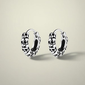 Op de afbeelding: Een paar zilveren oorringen met een schedelontwerp. De oorbellen hebben een gedetailleerd patroon van schedels en andere vormen. De binnenkant van de ringen is zwart, wat een contrast creëert met de zilveren buitenkant. De achtergrond is neutraal.