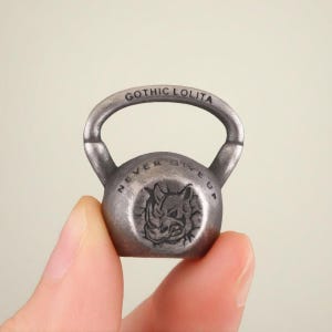 May include: A small, silver-coloured kettlebell with the words "GOTHIC LOLITA" arched across the top handle. The kettlebell has a circular emblem with a rhino head and the words "NEVER GIVE UP".