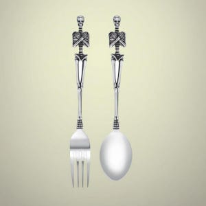Op de afbeelding: Een zilverkleurige vork- en lepelset. De handvatten zijn ontworpen met een skeletfiguur met gekruiste armen. De vork heeft vier tanden en de lepel heeft een afgeronde kom. De set is waarschijnlijk om te serveren.