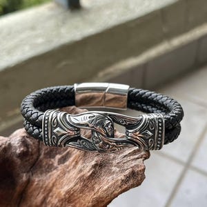 Puede incluir: Pulsera de cuero trenzado negro con cierre plateado. El cierre presenta un diseño ornamentado con detalles cobrizos. La pulsera se exhibe sobre un trozo de madera.