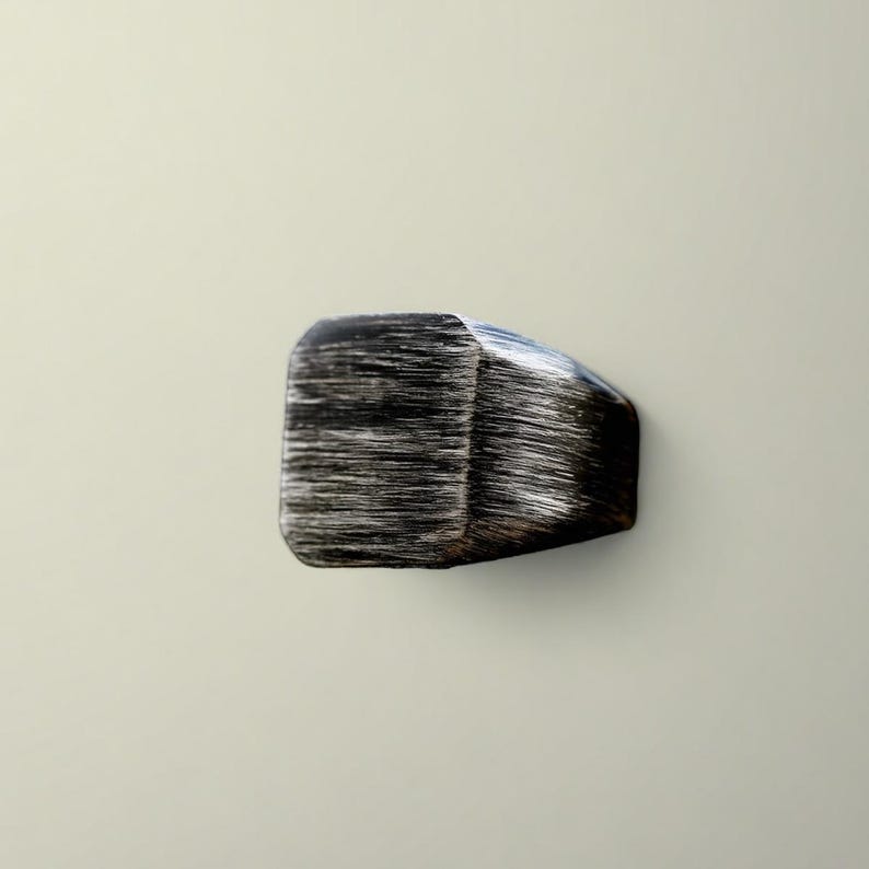 May include: A dark, rectangular cabinet knob with a textured, brushed metal appearance. The knob has a faceted design on one side and a rounded, smooth edge on the other. The color is primarily black and silver.