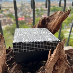 May include: A rectangular black box with a geometric patterned lid in shades of gray and white. The box is set on a piece of weathered, dark brown wood, with a blurred cityscape in the background.