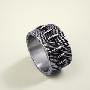 Pode incluir: Um anel prateado com uma superf&iacute;cie texturizada e um design costurado. O anel tem uma banda larga com linhas horizontais e detalhes verticais em forma de pontos. O anel &eacute; feito de metal e tem um acabamento fosco.
