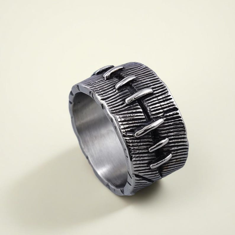 Pode incluir: Um anel prateado com uma superf&iacute;cie texturizada e um design costurado. O anel tem uma banda larga com linhas horizontais e detalhes verticais em forma de pontos. O anel &eacute; feito de metal e tem um acabamento fosco.