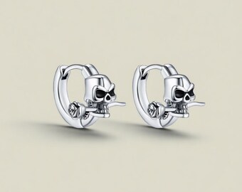 May include: A pair of silver hoop earrings. Each earring features a skull with black eyes and a rose. The skull is holding a small object in its mouth. The earrings are displayed against a neutral background.