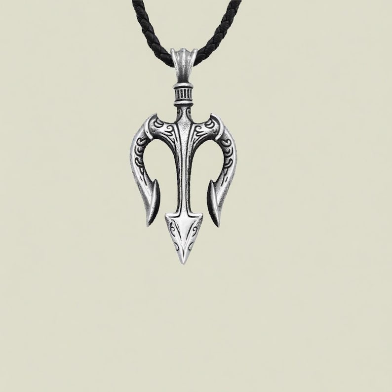May include: Silver trident pendant with detailed patterns, hanging from a black braided cord. The pendant has a central spearhead design with curved prongs, giving it a symmetrical and decorative look.