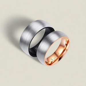 Puede incluir: Dos anillos de metal cepillado. Uno es plateado con interior negro. El otro es plateado con interior oro rosa. El anillo de oro rosa tiene el texto "S. steel" grabado en el interior.