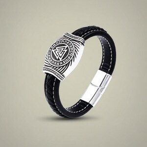Pode incluir: Pulseira de couro preta com fecho prateado e peça central decorativa. A peça central apresenta um design detalhado com um símbolo triangular e inscrições rúnicas. A pulseira tem um design trançado e costuras brancas.