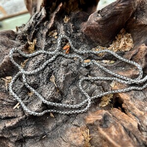 May include: A silver chain necklace with a box chain design, resting on a piece of weathered, dark brown wood. The necklace is made of small, square links, creating a textured look. The wood has a rough, natural texture.