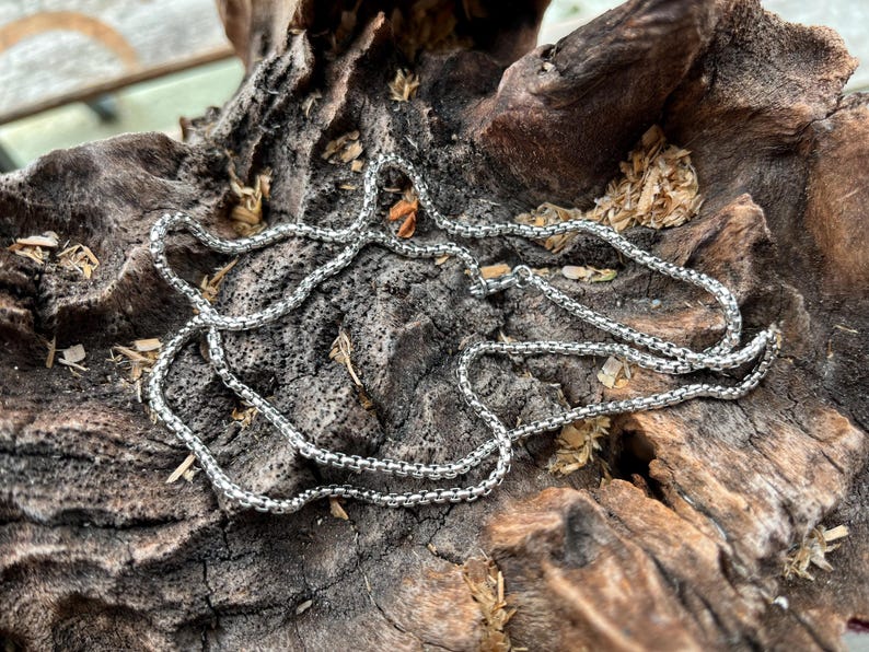 May include: A silver chain necklace with a box chain design, resting on a piece of weathered, dark brown wood. The necklace is made of small, square links, creating a textured look. The wood has a rough, natural texture.