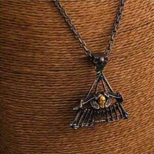 Puede incluir: Un collar con colgante plateado con un diseño triangular, que presenta un motivo de calavera y huesos. El colgante tiene una calavera de color dorado en el centro. El collar se muestra sobre un fondo marrón.