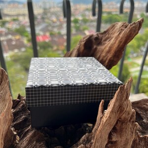 May include: A rectangular black box with a patterned lid featuring a repeating geometric design in shades of gray and white. The box is set against a natural wood background, with a city view in the distance.