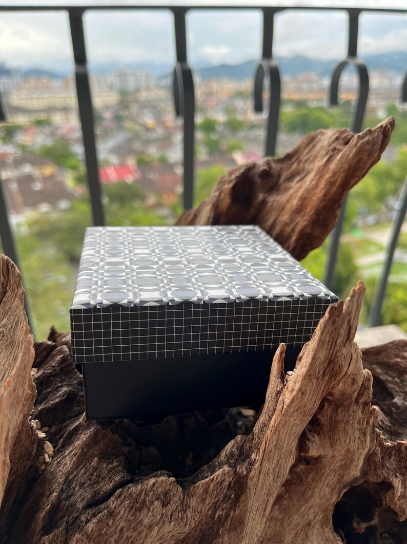 May include: A rectangular black box with a patterned lid featuring a repeating geometric design in shades of gray and white. The box is set against a natural wood background, with a city view in the distance.