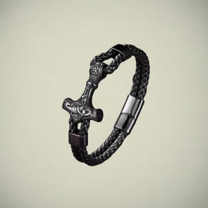 Puede incluir: Pulsera de cuero trenzado negro con un colgante detallado de martillo de Thor en tono plateado. La pulsera tiene un cierre magnético y está diseñada con una temática vikinga. El cuero parece tener aproximadamente 1,3 cm de ancho.