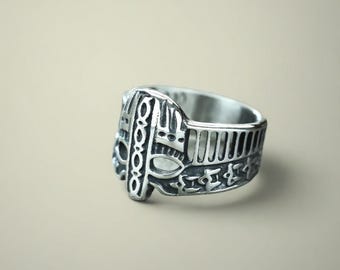 May include: A silver ring with an elaborate design. The ring showcases a detailed pattern of vertical lines, geometric shapes, and stylized symbols. The ring is likely made of metal and has a polished finish.