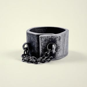Op de afbeelding: Een verweerde metalen enkelband met een geklonken sluiting en een korte ketting. De enkelband is cilindrisch van vorm met een donkere, verouderde afwerking, wat wijst op een vintage of antieke uitstraling. De ketting bestaat uit kleine schakels.
