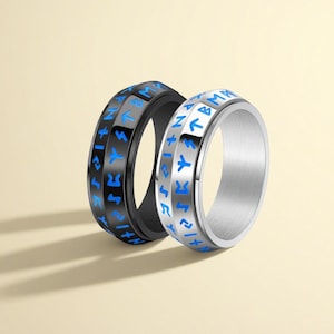 Op de afbeelding: Twee roestvrijstalen ringen met blauw gegraveerde symbolen. Eén ring is zwart, de andere is zilver. De symbolen zijn gelijkmatig verdeeld over de buitenband van de ringen. De ringen worden weergegeven op een lichtbeige achtergrond.
