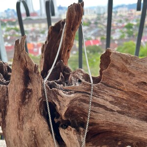 May include: A silver box chain necklace is presented against a backdrop of textured, brown wood. The necklace hangs vertically, highlighting its delicate links. The background features a blurred cityscape.