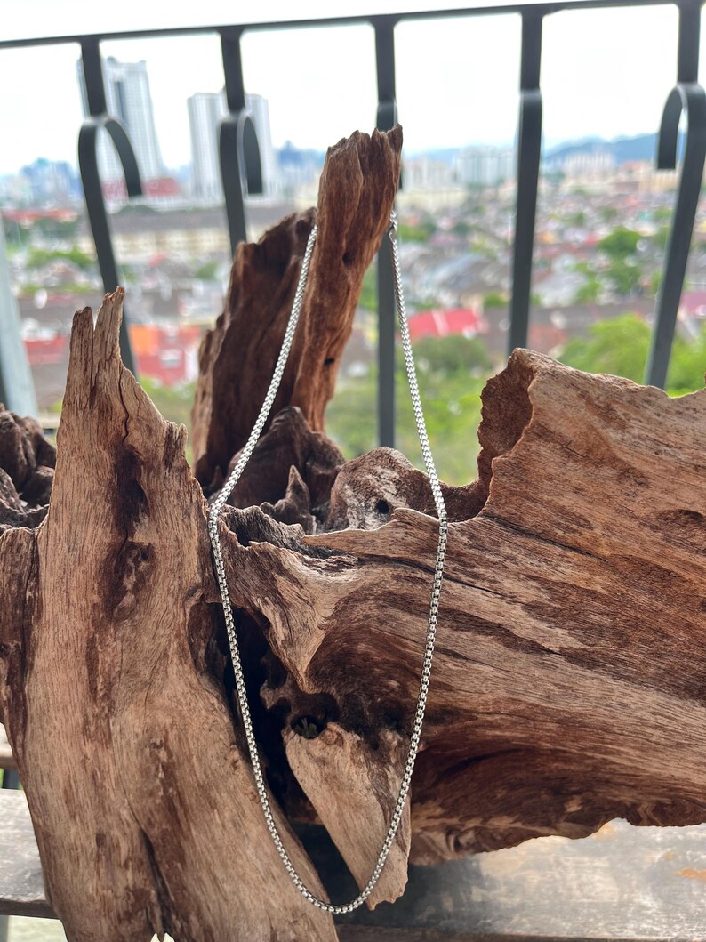 May include: A silver box chain necklace is presented against a backdrop of textured, brown wood. The necklace hangs vertically, highlighting its delicate links. The background features a blurred cityscape.