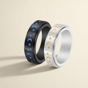 Könnte beinhalten: Zwei Spinner-Ringe mit Himmelsmotiven. Ein Ring ist schwarz mit blauen Monden und Sternen, der andere silberfarben mit goldenen Monden und Sternen. Die Ringe sind zum Drehen konzipiert und bieten ein taktiles Merkmal.