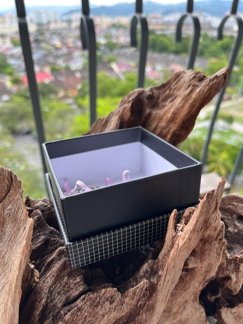 May include: A black gift box with a white interior sits on a patterned box, placed on driftwood. The box contains small, light purple objects. The background shows a blurred cityscape and a decorative metal railing.