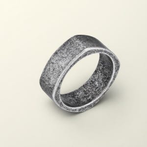 Puede incluir: Un anillo plateado texturizado con forma rectangular. El anillo tiene un aspecto desgastado y antiguo, con una textura superficial rugosa. El diseño del anillo es simple y moderno, adecuado para el uso diario.