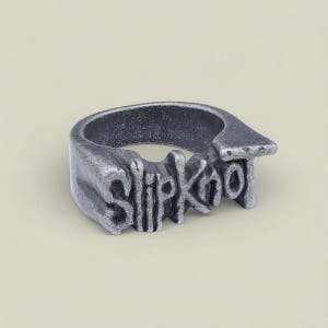 May include: A silver ring featuring the band name "Slipknot" in a distinctive font. The ring has a textured, aged look, possibly with an industrial design. A statement piece of jewellery, it is likely made of metal.