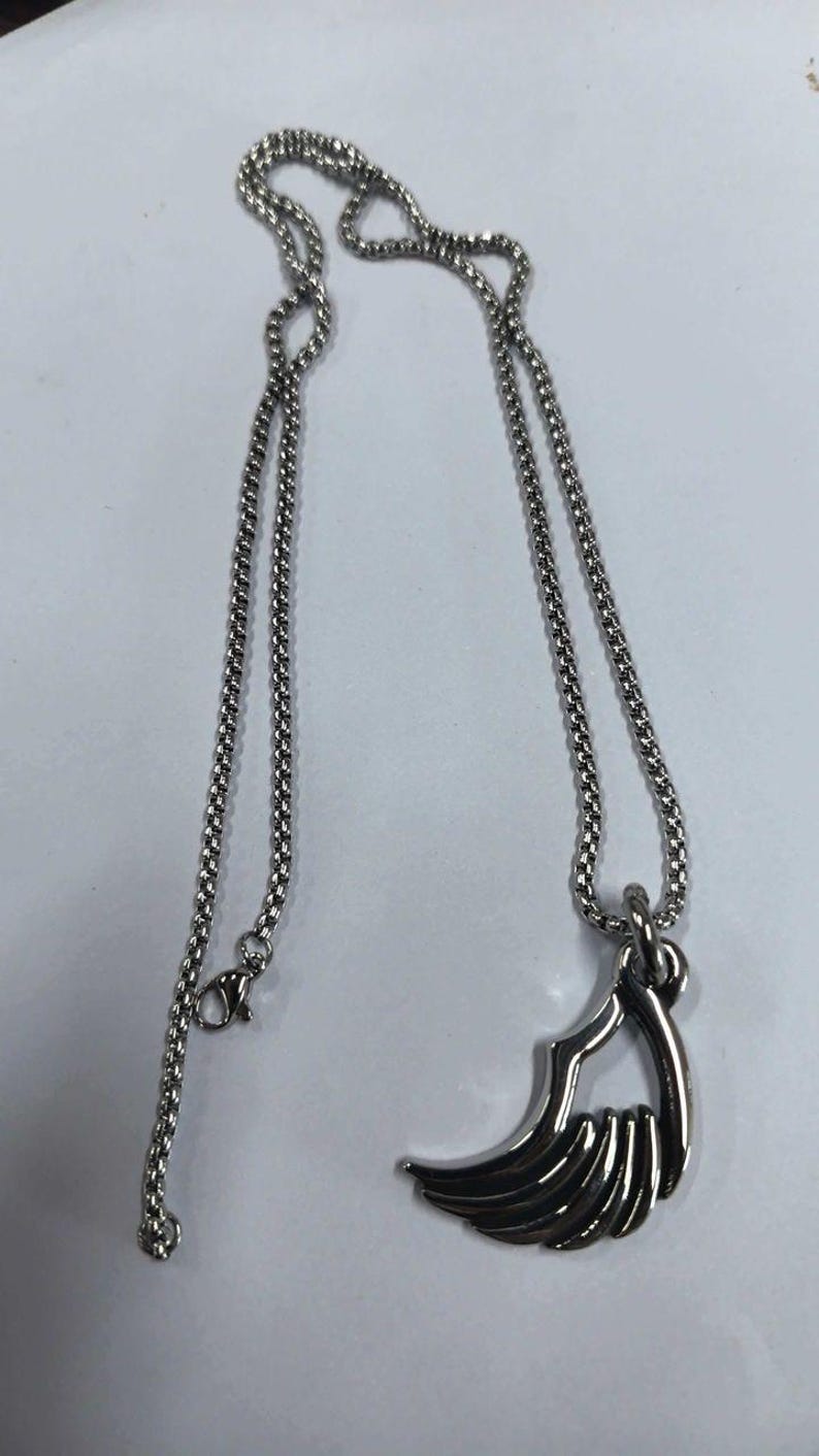 May include: A silver-colored necklace with a pendant shaped like a stylized wing. The chain is a fine, linked design, and the pendant has a detailed, layered appearance. The necklace is displayed on a white surface.