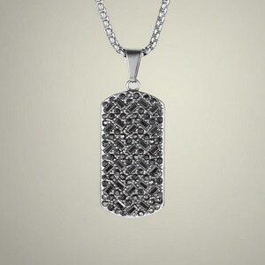 Puede incluir: Collar plateado con un colgante rectangular. El colgante está cubierto de pequeñas piedras de color gris oscuro dispuestas en un patrón geométrico. La cadena es de tipo caja.