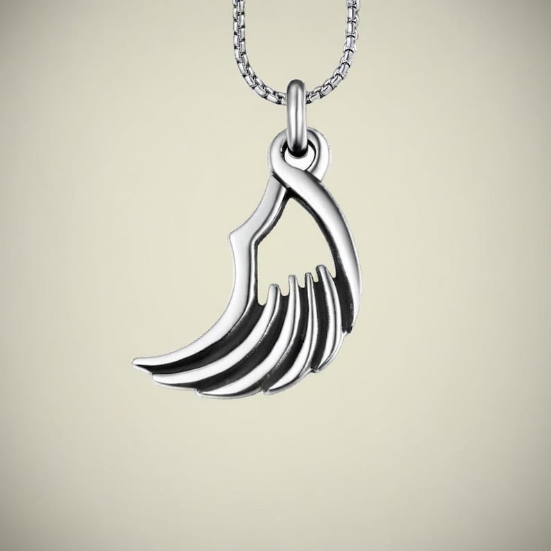 May include: A silver-colored pendant necklace featuring a stylized wing design. The pendant has a curved shape with multiple parallel lines, suspended from a delicate chain. The necklace is displayed against a soft, neutral background.