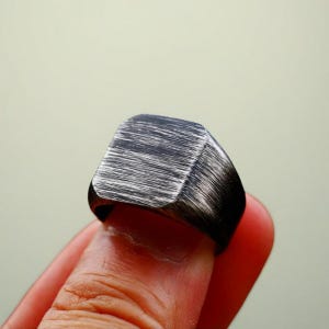 May include: A black and silver-toned ring with a square face. The ring has a brushed metal texture, with visible horizontal lines. The ring is worn on a finger, showcasing its design and craftsmanship. A piece of jewelry.