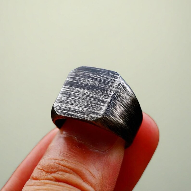 May include: A black and silver-toned ring with a square face. The ring has a brushed metal texture, with visible horizontal lines. The ring is worn on a finger, showcasing its design and craftsmanship. A piece of jewelry.
