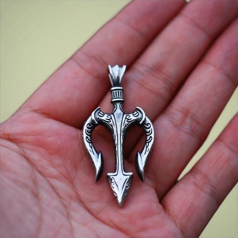 May include: A silver trident pendant with detailed patterns. The pendant has a central spearhead design with two curved prongs. The top has a loop for a chain. The pendant is held in a hand. The pendant is approximately 6 cm in length.
