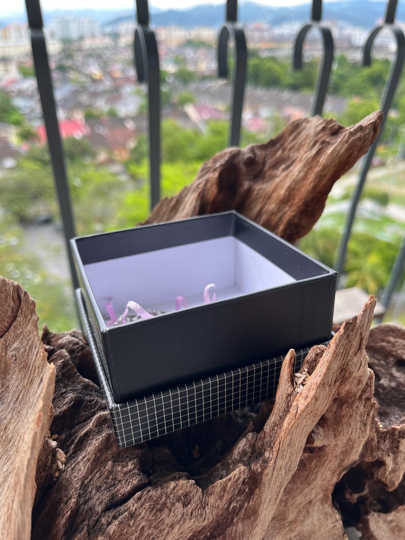 May include: A black gift box with a white interior sits atop a patterned box, resting on a piece of driftwood. The box contains small, light purple objects. The background features a blurred view of a cityscape and a decorative metal railing.